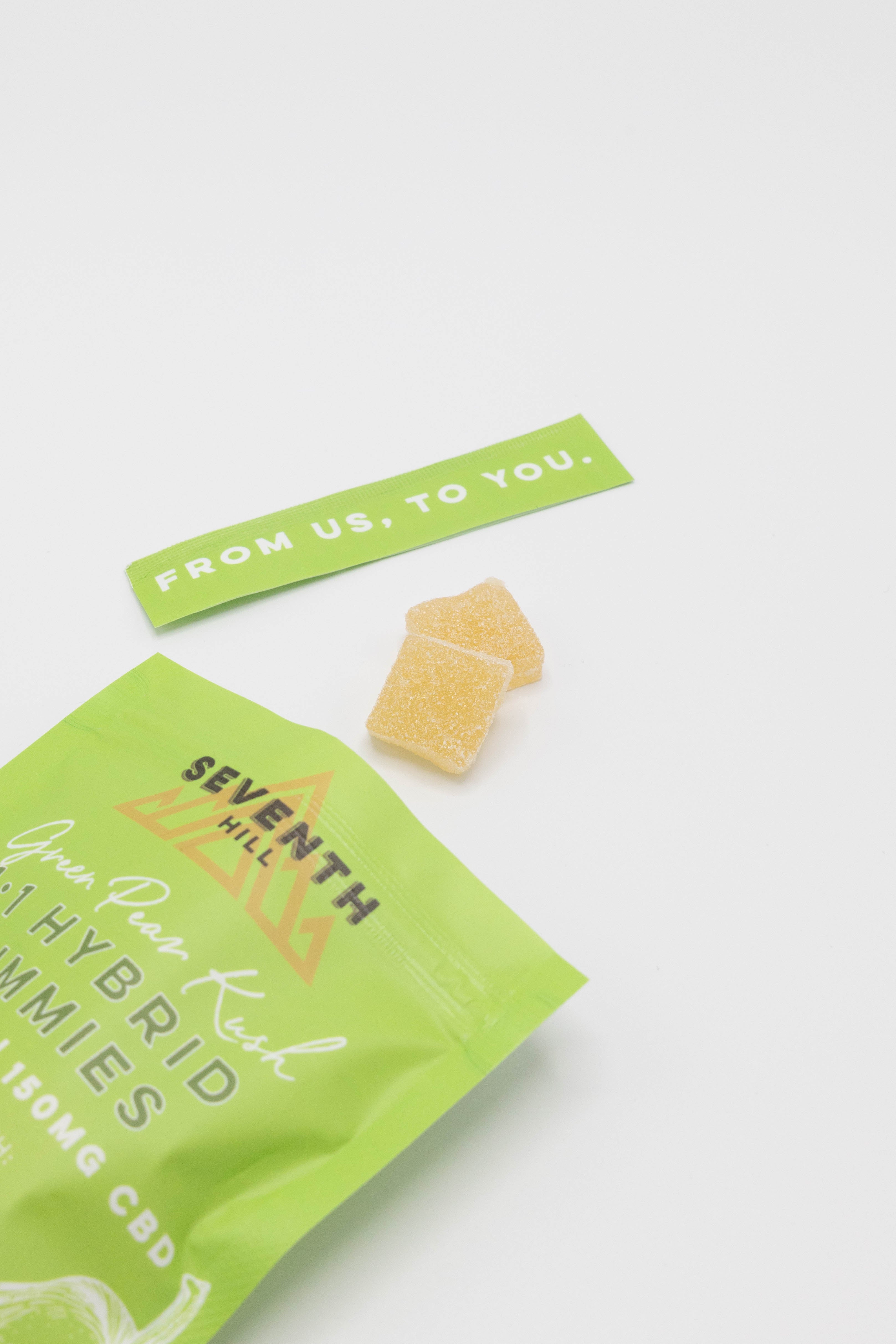 A bright green bag of Seventh Hill gummies with two small yellow gummy candies and a green paper tag reading 'FROM US, TO YOU.' on a clean white background.