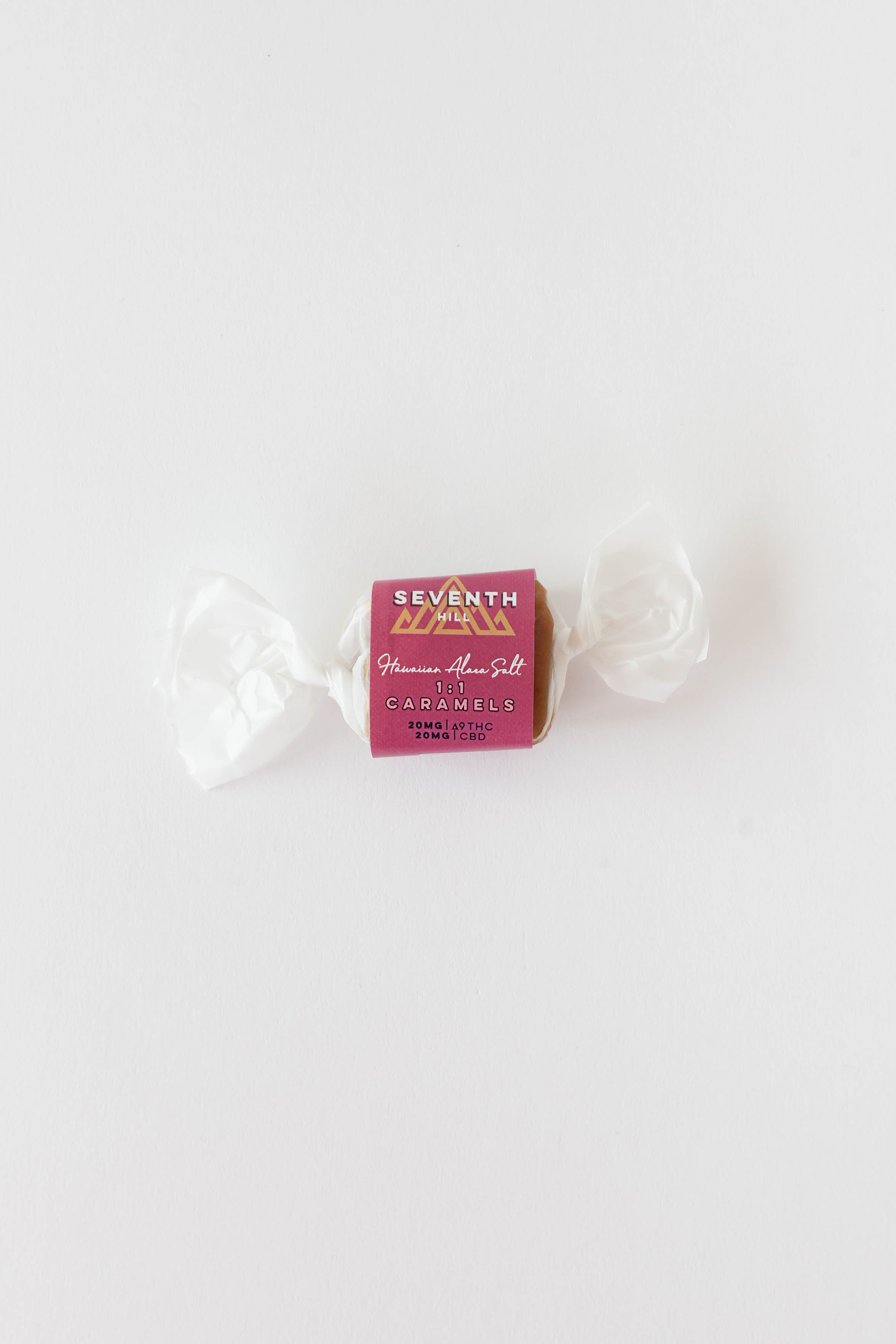 A single wrapped caramel candy from Seventh Hill, featuring a magenta wrapper with gold lettering and white wax twist ties, set against a plain white background.