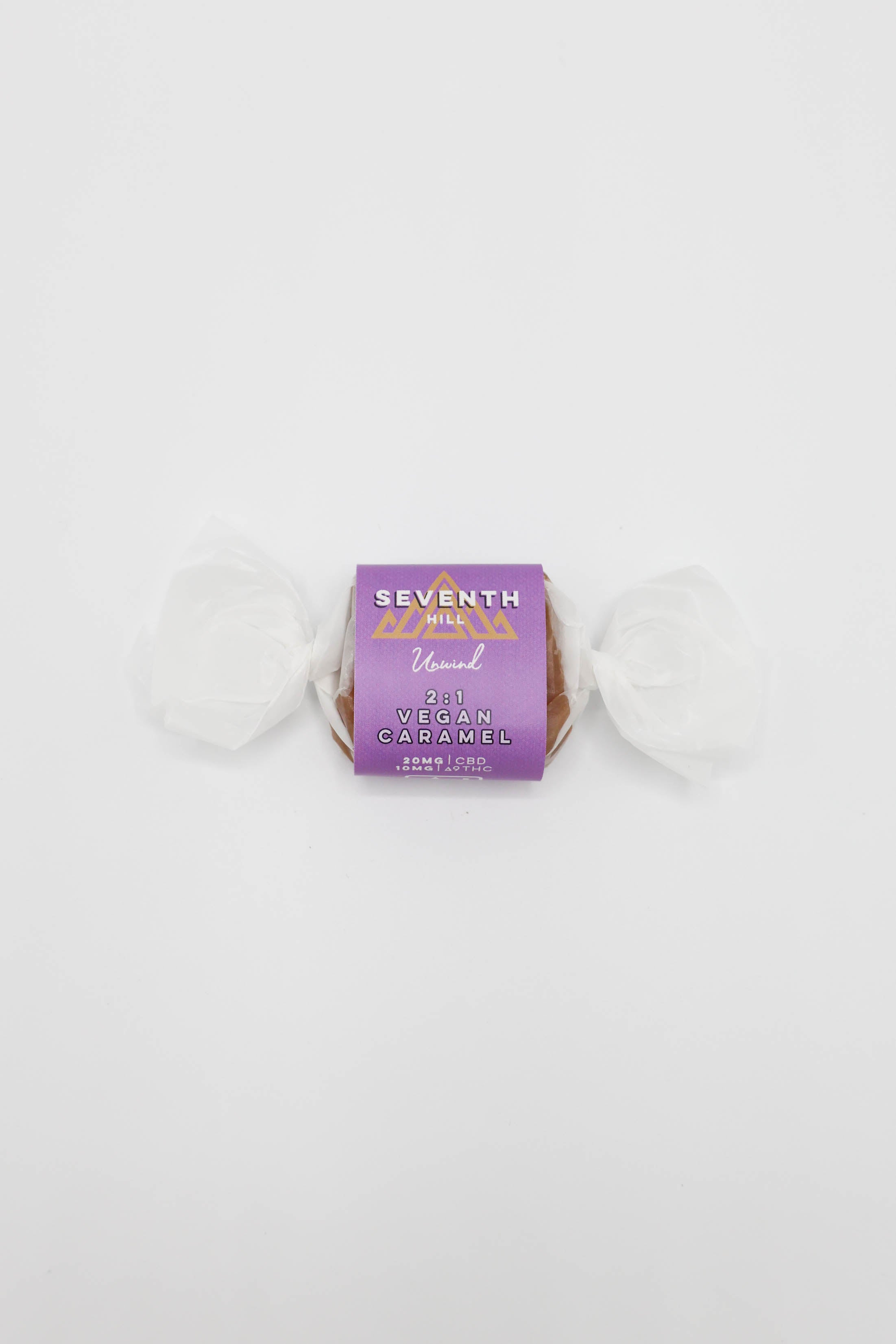 A single wrapped vegan caramel candy from the brand Seventh Hill, featuring a purple wrapper with gold lettering and white wax paper ends, set against a plain white background.