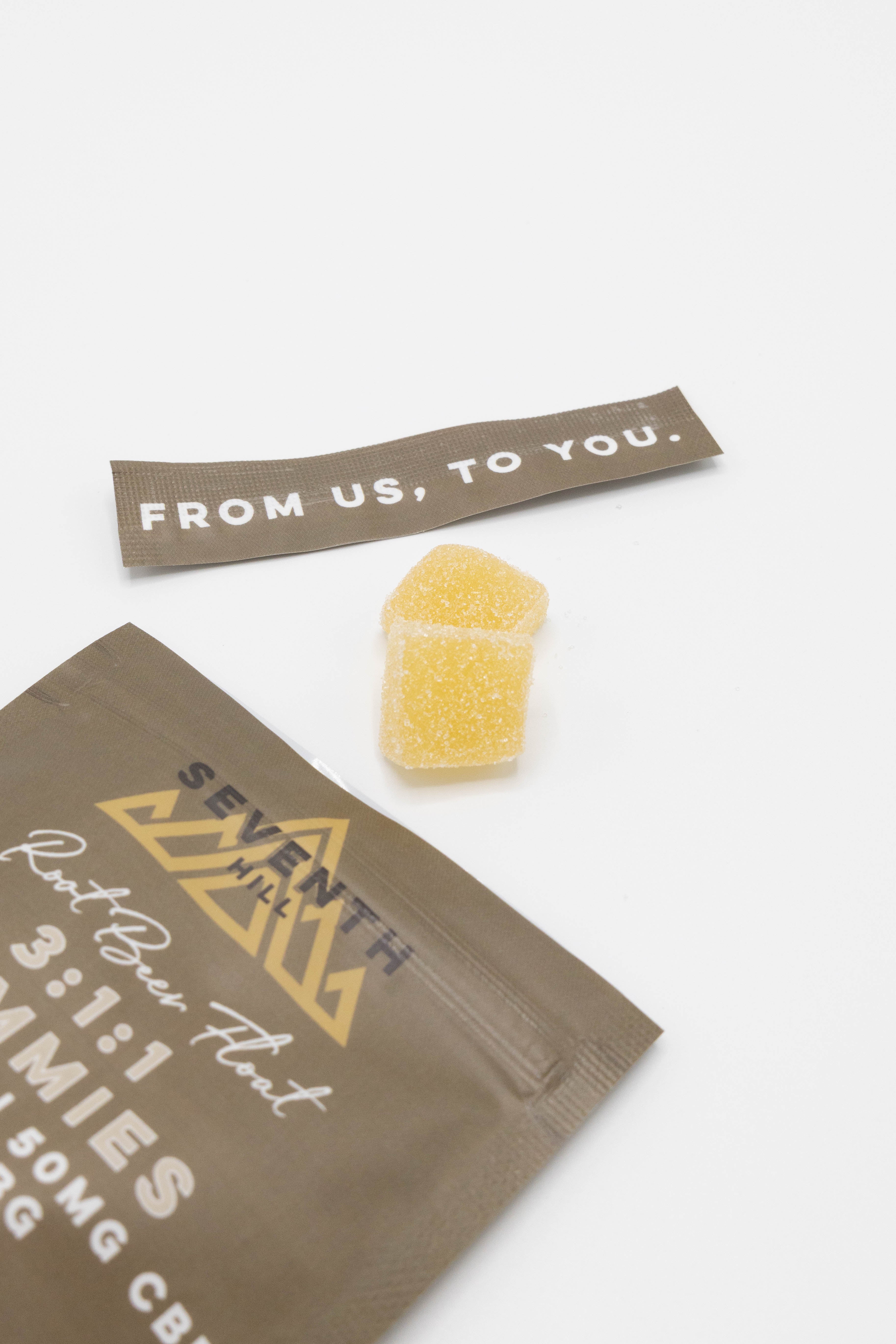 A close-up of a Seventh Hill gummy package in brown with gold and white lettering, alongside a small yellow gummy candy and a brown tag that reads 'FROM US, TO YOU.' on a clean white background.
