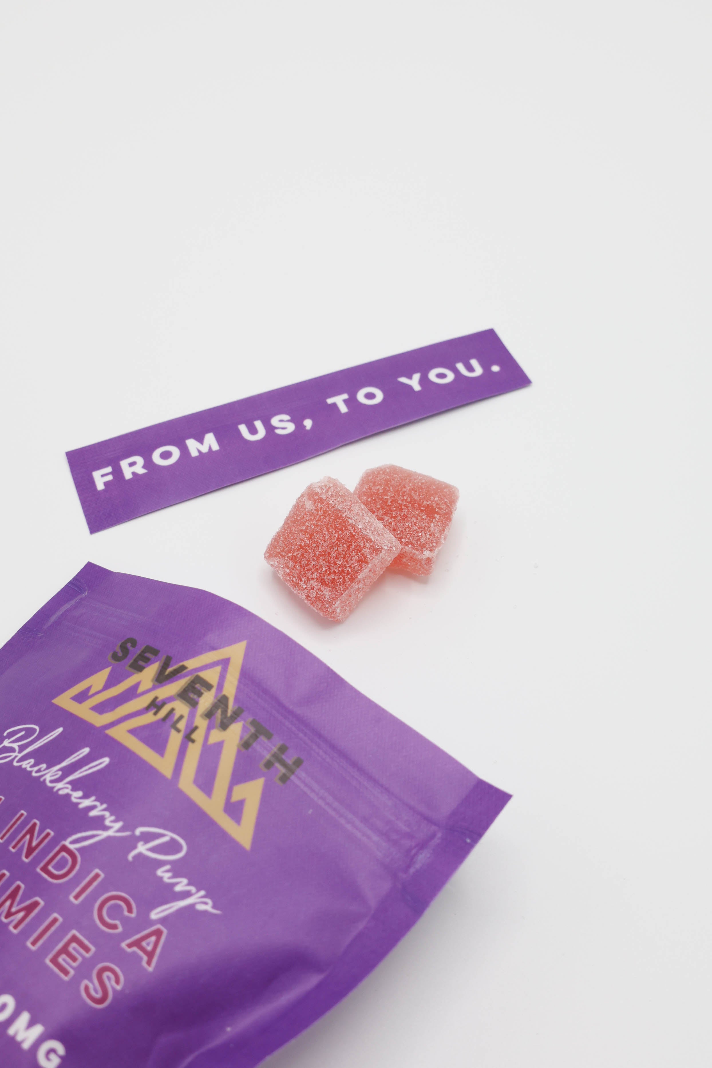 Purple bag of Seventh Age Blackberry Purp 3:1 Indica candy with two pink gummy candies and a purple tag reading 'FROM US, TO YOU.' on a white background.