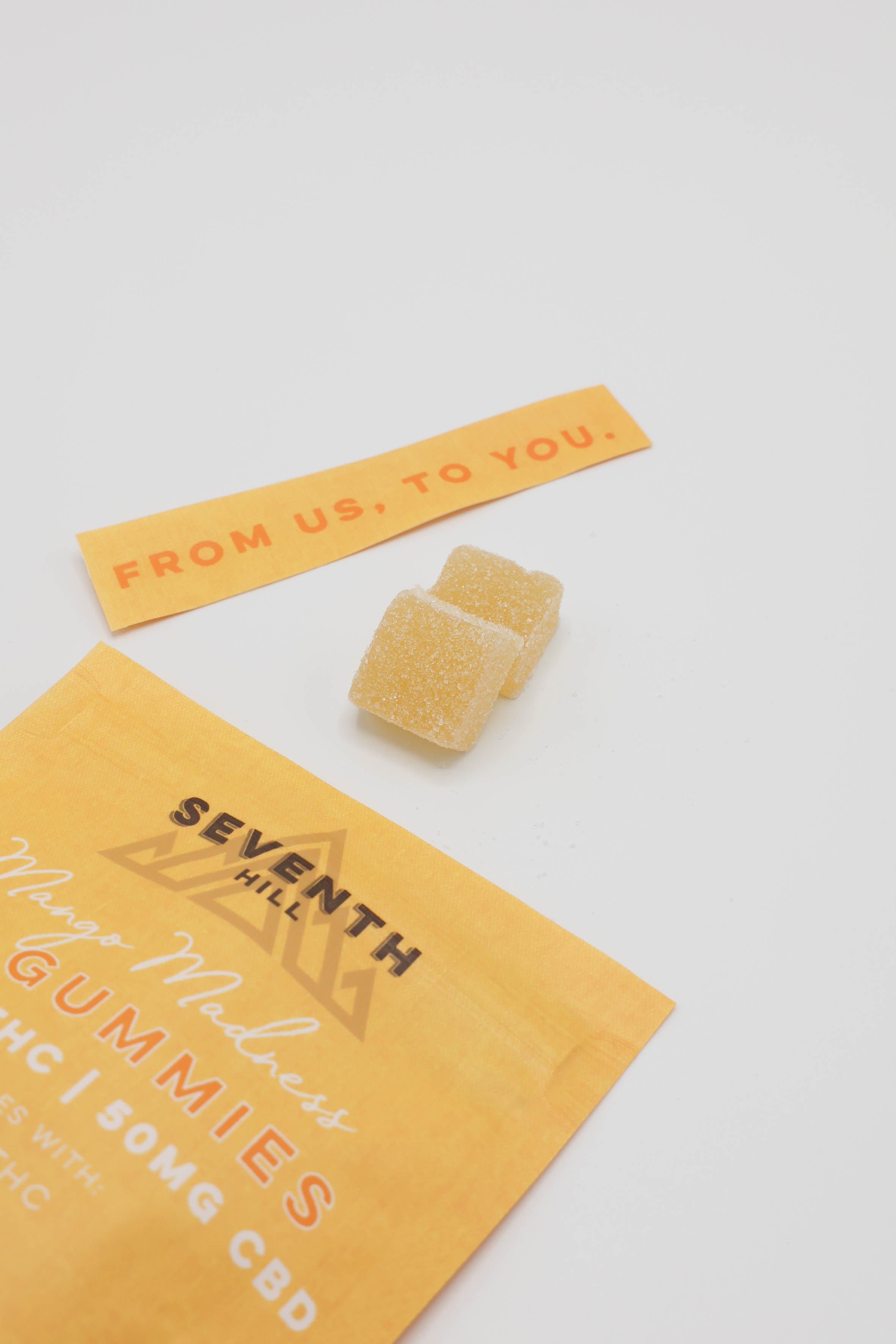 Two yellow gummy candies and a yellow paper sleeve of Seventh Hill Mango Madness 3:1 Gummies on a white background.