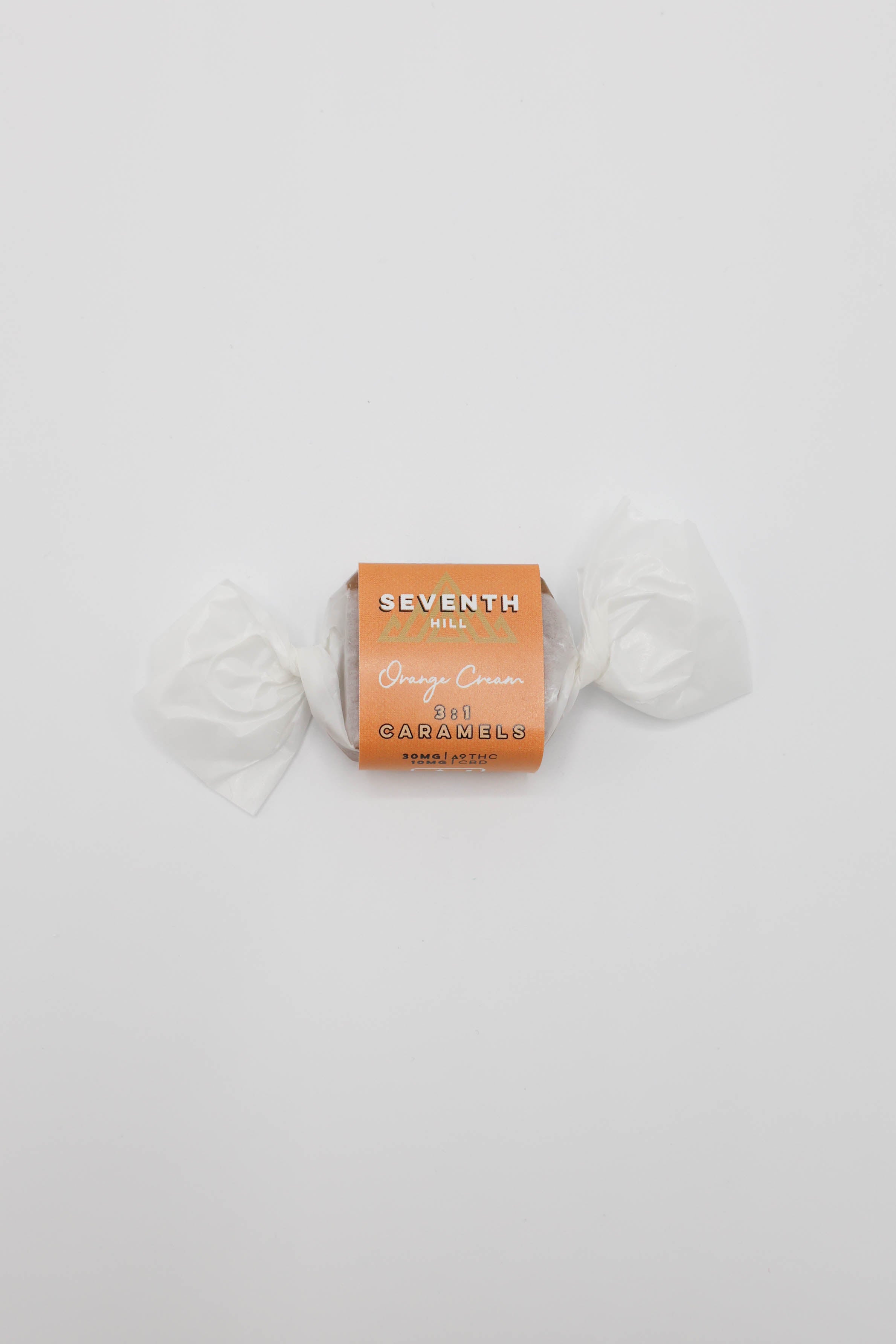 A wrapped piece of candy from Seventh Hill, specifically 3:1 Orange Cream Caramel, wrapped in white paper with a tan-orange cardboard label, centered on a plain white background.