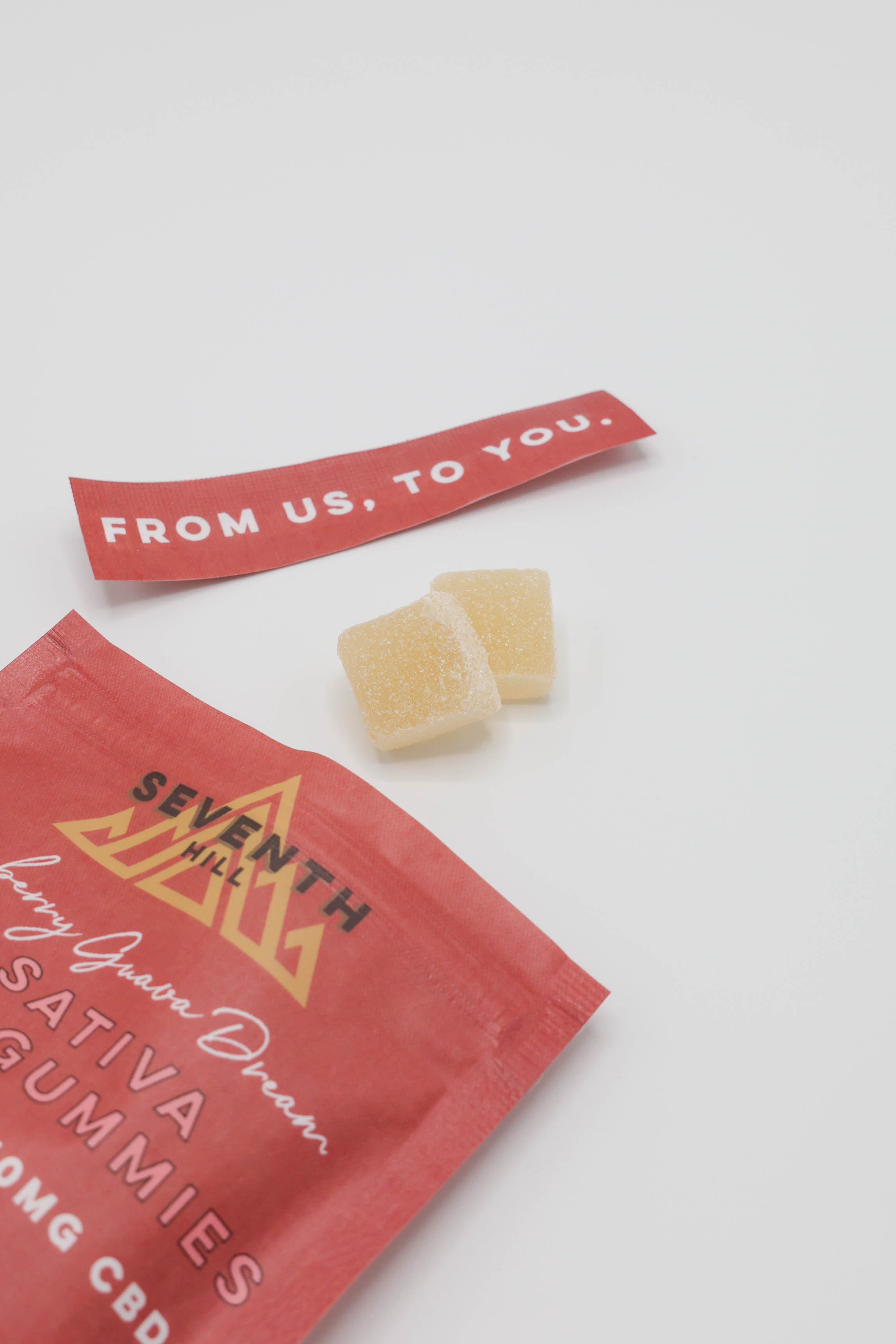 A red packet of Seventh Hill 3:1 Sativa Hybrid Gummies with two pale yellow gummy cubes placed on top, alongside a red tag reading 'FROM US, TO YOU.' on a clean white background.