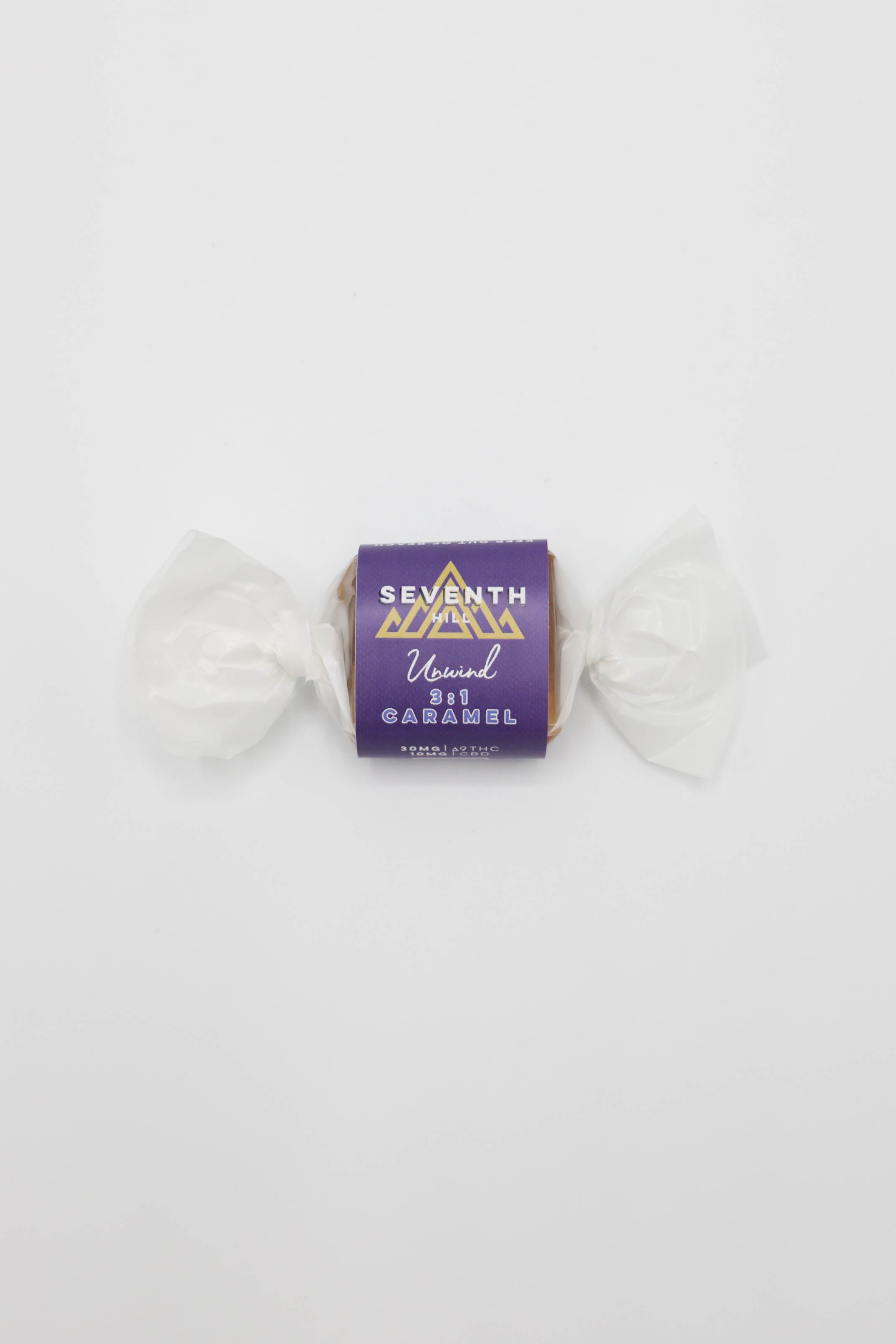 A single wrapped candy on a plain white background. The candy is wrapped in white wax paper with a purple label in the center that reads 'SEVENTH HILL 3:1 Unwind Caramel"