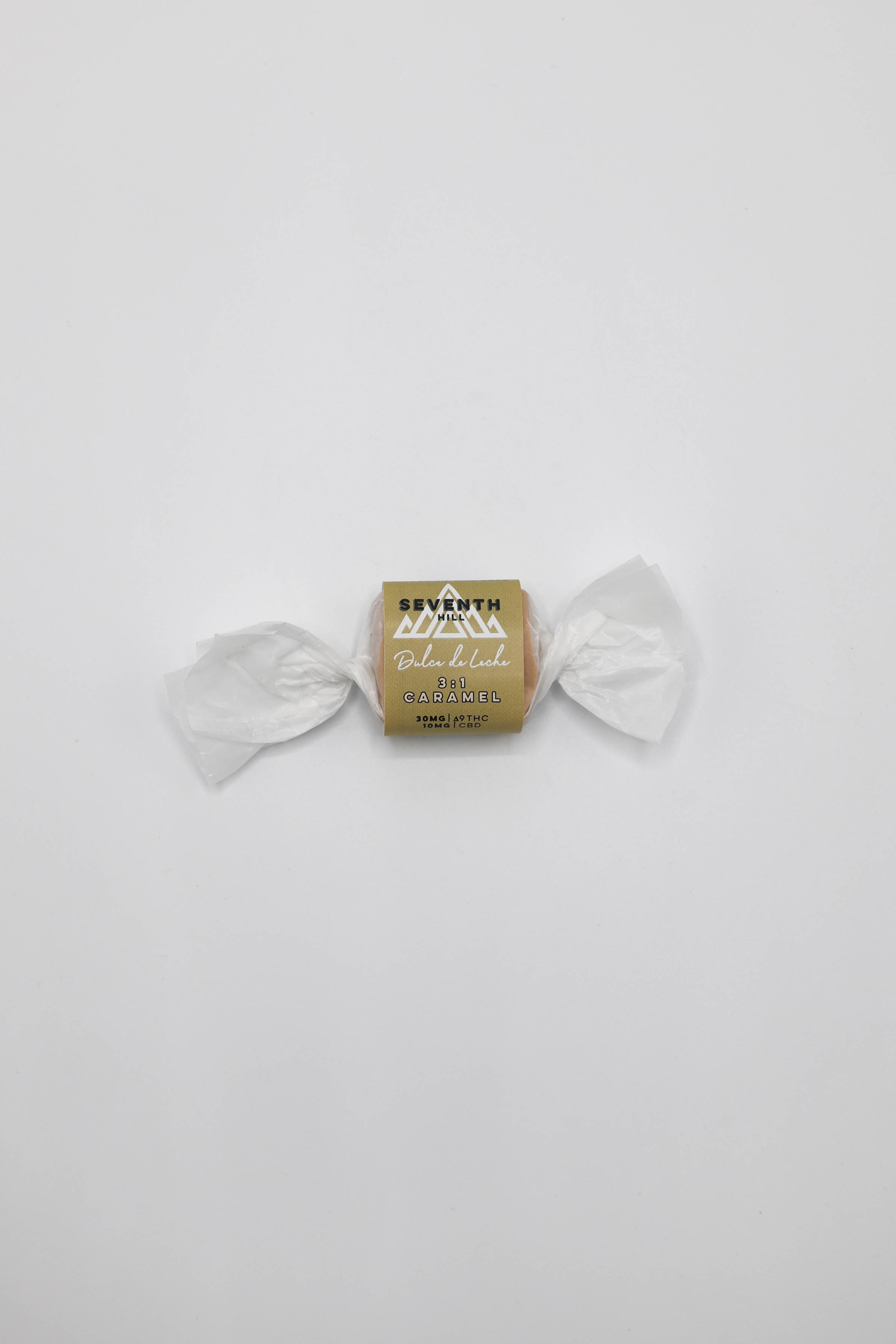 A single wrapped candy centered on a plain light gray background. The center wrapper is tan/gold with black and white text reading 'SEVENTH Hill' and 'Dulce de Leche'.