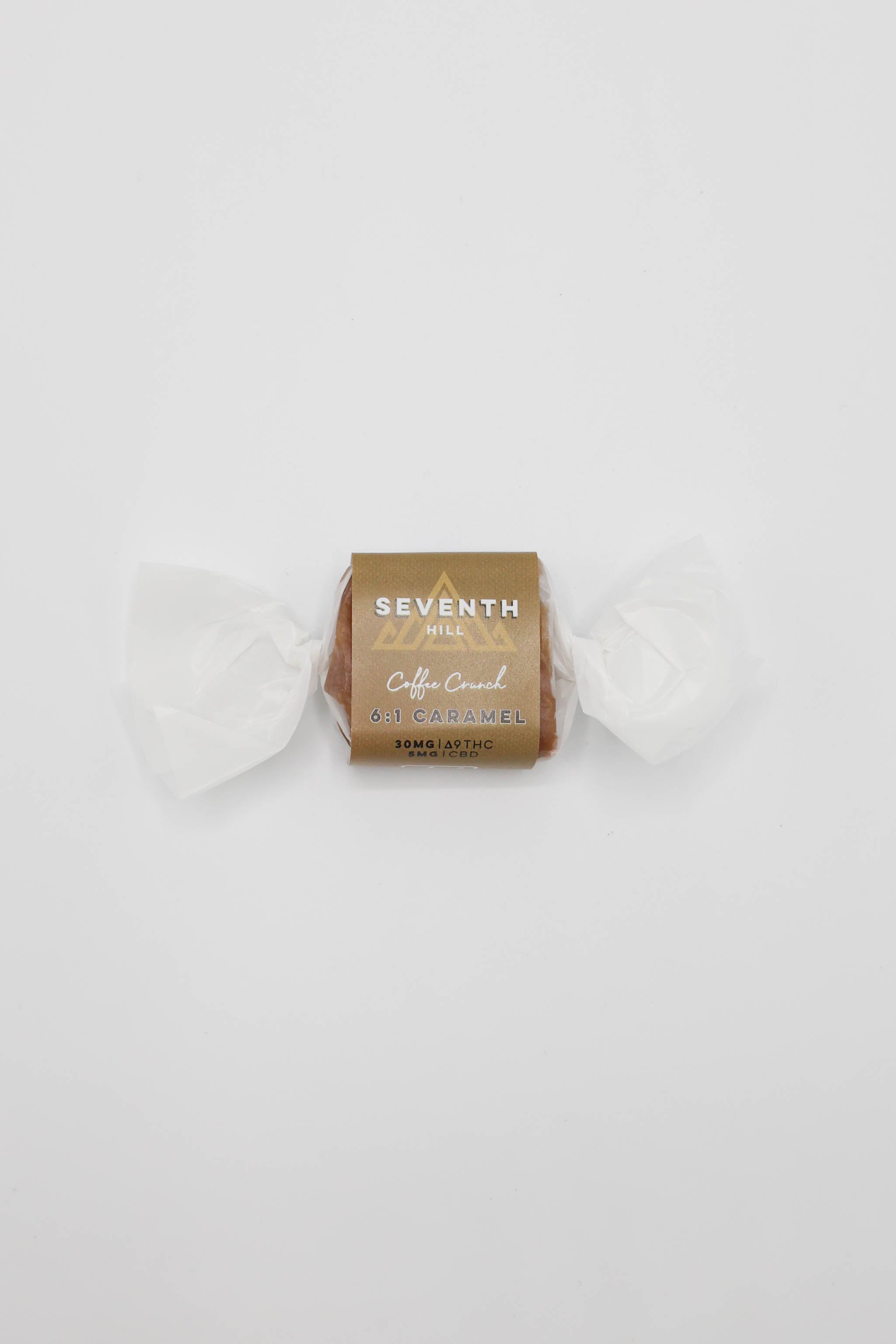 A single wrapped piece of caramel from the brand Seventh Hill, specifically 6:1 Coffee Crunch, wrapped in white wax paper and a brown paper sleeve, centered on a plain white background.