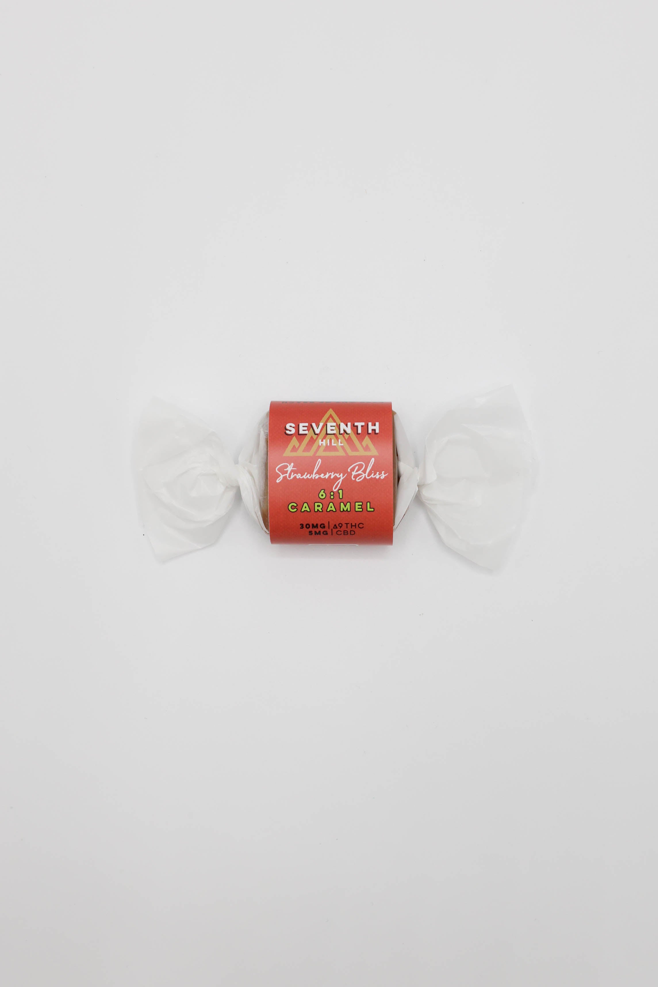 A single wrapped piece of caramel on a plain white background. The wrapper is red with white text reading 'SEVENTH HILL' and 'Strawberry Bliss', and the candy is wrapped in white wax paper with white paper ties on each side.