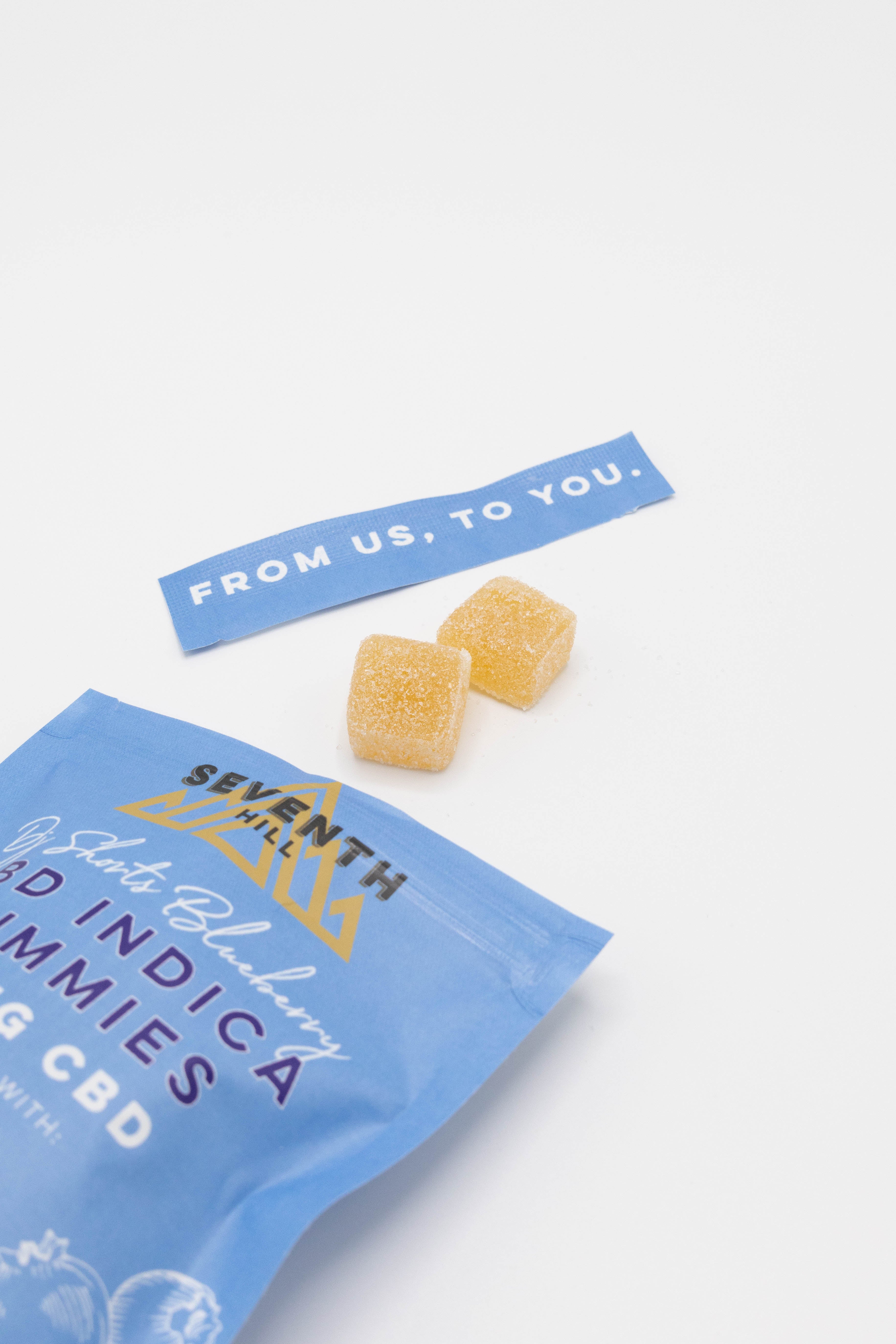 A blue bag of Seventh Hill CBD gummies lies on a white surface with two small yellow-orange gummy cubes placed above it and a blue tag reading 'FROM US, TO YOU.'.