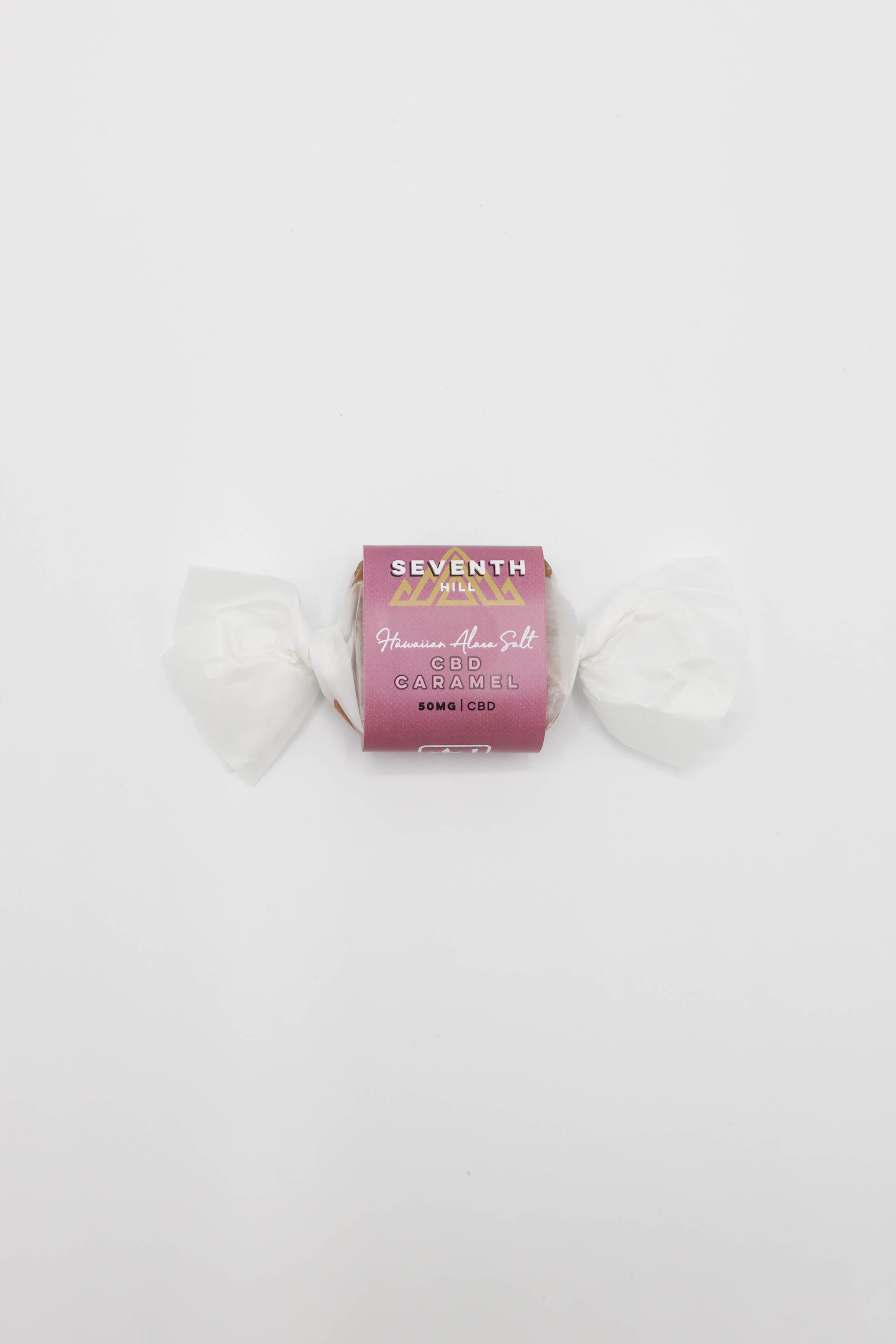 A single wrapped piece of candy on a plain white background. The wrapper is dark pink/magenta with gold and white text reading 'SEVENTH HILL' and 'CBD Hawaiian Alaea Salt Caramel'.