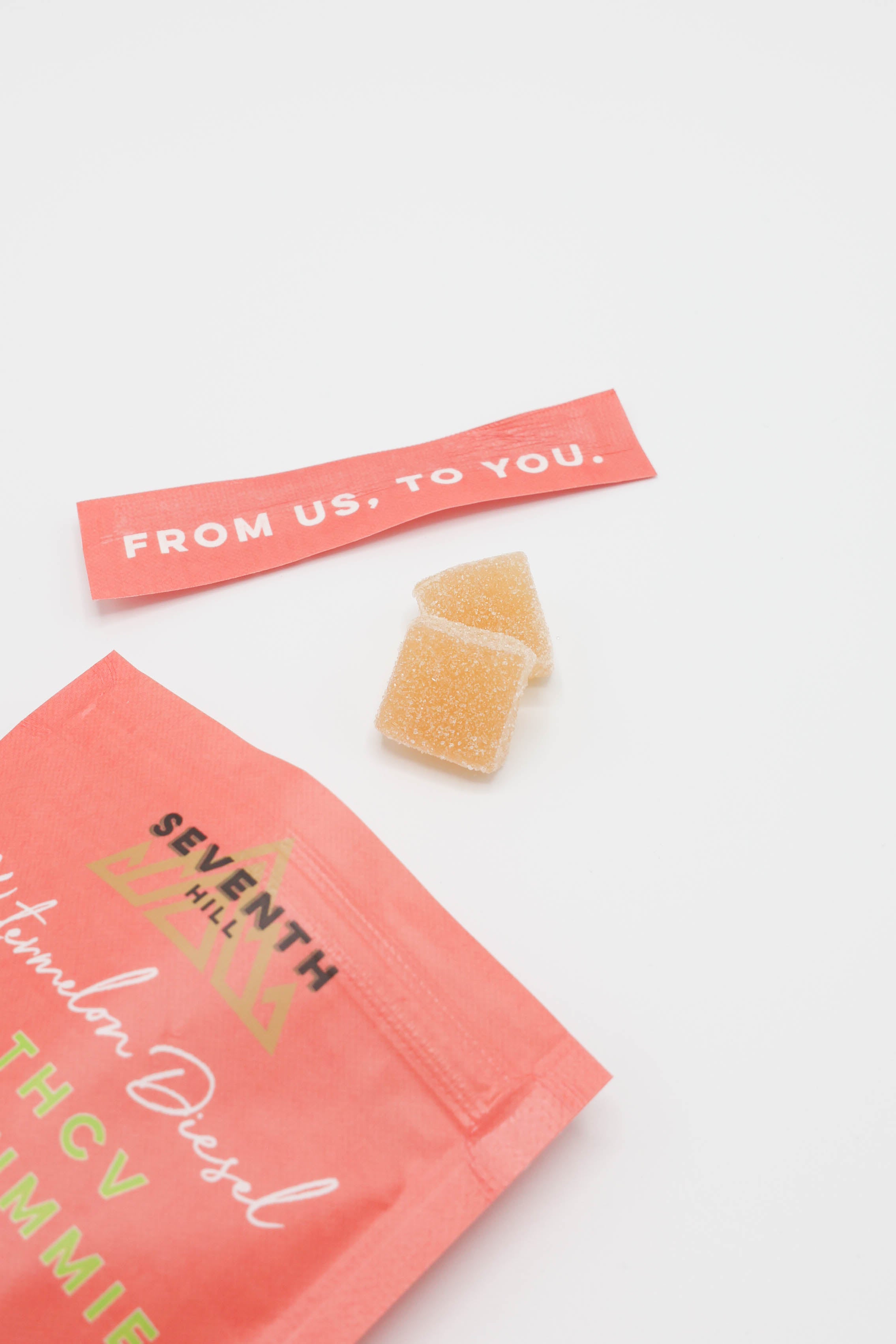 A coral pink bag of Seventh Hill THCV gummies with a matching paper tag reading 'FROM US, TO YOU.' and two translucent orange gummies on a clean white background.